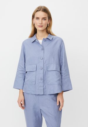 Chaqueta de lino azul claro con cuello, cierre frontal con botones y dos grandes bolsillos exteriores. Presenta mangas tres cuartos y un ajuste relajado.