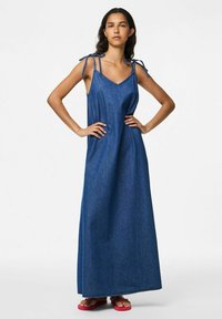 Denim maksi haljina s dubokim V izrezom, podesivim vezicama i slobodnim krojem, u srednje plavoj boji. Teksturična tkanina s bočnim džepovima.