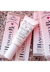 Too Faced Hangover RX face primer in a white tube with gold accents, surrounded by pink cardboard boxes featuring similar design elements.