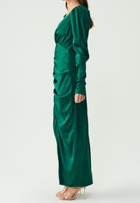 Grön satin klänning med långa puffärmar och djup V-ringning. Har rynkade detaljer vid midjan och en rak kjol. Klara klackar visas.