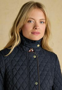 Námornícky prešívaný kabát s vysokým golierom, s diamantovými vzormi, cvočkovými gombíkmi a hladkou textúrou. Doplnkovaný červenou podšívkou.