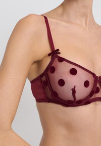 Soutien-gorge bordeaux en tulle transparent avec des pois en velours. Il présente des fines bretelles et de petits nœuds aux jonctions des bretelles. Texture lisse.