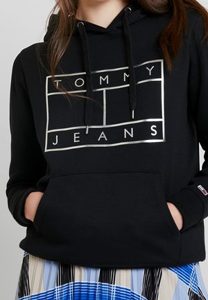 Osoba nosząca czarną bluzę z kapturem z logo "TOMMY JEANS" w białych obrysowanych ramkach oraz z rękami w przedniej kieszeni, w połączeniu z wzorzystą spódnicą.