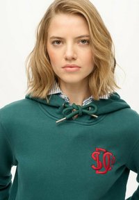Ung kvinde med skulderlangt blond hår iført en grøn hoodie med et rødt broderet logo og en stribet skjorte med krave under.