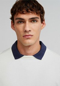 Cămașă polo albă cu mâneci lungi și guler bleumarin. Textură netedă, design simplu și gulerul în contrast evidențiază decolteul.
