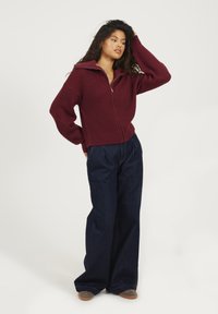 Burgundowy sweter zapinany na zamek z wysokim kołnierzem, w połączeniu z luźnymi ciemnoniebieskimi spodniami dżinsowymi i jasnobrązowym obuwiem. Sweter ma prążkowaną teksturę.