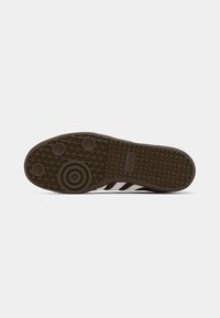 Sål af mørkebrune Adidas-sneakers med cirkulære og trekantede dækmønstre og synlige tre hvide striber på siden.
