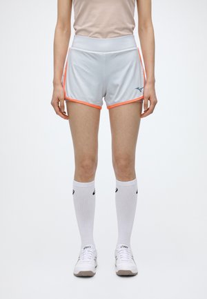 Un mannequin porte un short de course blanc à bordure corail, des chaussettes blanches montant jusqu’aux genoux et des chaussures de course blanches. Il se tient devant un fond uni.