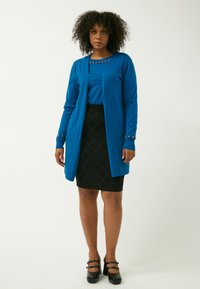 Blauer Cardigan mit langen Ärmeln, kombiniert mit einem blauen Oberteil und einem schwarzen Bleistiftrock mit Diamantmuster, getragen mit schwarzen hohen Schuhen.