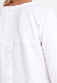 White top with a contrasting textured panel, featuring a round neckline and button closures down the back. Smooth fabric with a subtle sheen.