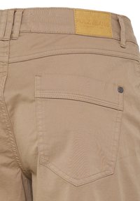 Beżowe spodnie pokazujące tylną kieszeń oraz brązową skórzaną metkę z napisem "Pulz Jeans" powyżej pasa.