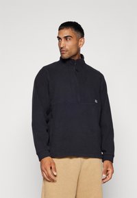 Granatowy polarowy sweter z wysokim kołnierzem, na pół zamkiem i detalami szwów. Miękka tekstura i luźny krój. Noszony z beżowymi spodniami.