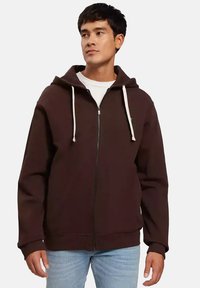 Brązowa bluza z kapturem na zamek błyskawiczny, z regulowanym kapturem, z przodu na zamek oraz ściągaczami przy mankietach i u dołu. Nosi się ją na białej koszuli, w połączeniu z jasnymi dżinsami.