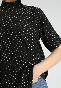 Svart kortärmad topp med hög krage, prydd med gula prickar, slät textur och en lös passform. Har sidfickor.