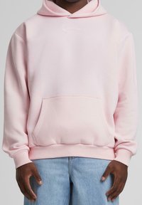 Man i ljusrosa hoodie med framficka och ljusblå jeans, stående med händerna avslappnat vid sidorna mot en neutral bakgrund.