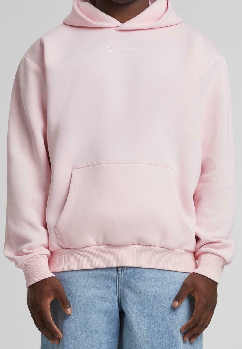 Man i ljusrosa hoodie med framficka och ljusblå jeans, stående med händerna avslappnat vid sidorna mot en neutral bakgrund.