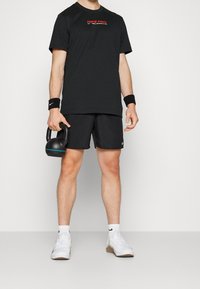 Mustat Nike Pro -t-paidat, joissa on punainen logo, yhdistettynä mustiinShortseihin, valkoisiin tossuihin ja ranteenauhoihin. Pidät mustaa kettlebelliä, jossa on sinisiä yksityiskohtia.