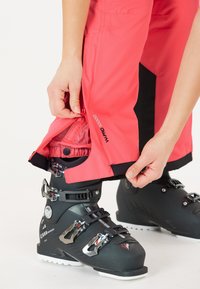 Roze skibroek met zwarte accenten, verstelbare boorden en ritssluitingen. Een zwarte skiboot is gedeeltelijk zichtbaar in het kader.