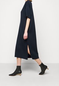 Personne marchant dans une robe midi bleu marine avec une fente latérale, portant des bottines noires à petits talons en bois sur un fond clair.