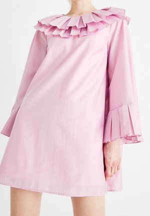 Frau in einem hellrosa Kleid mit mehrlagigen, plissierten Rüschen am Kragen und an den Ärmeln, von Hals bis Mitte Oberschenkel zu sehen.