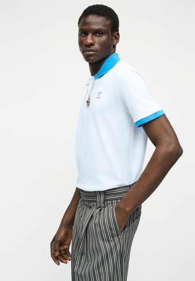 Άνδρας που φοράει λευκό πόλο με μπλε γιακά και ριγέ μαύρο-λευκό παντελόνι, στέκεται με το ένα χέρι στην τσέπη μπροστά σε απλό φόντο.