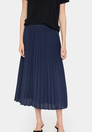 Femme portant une jupe midi plissée bleu marine associée à un top noir à manches courtes et des chaussures noires slip-on, debout sur un fond blanc.
