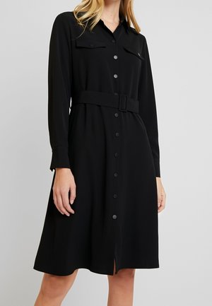 Kobieta ubrana w czarną sukienkę z długim rękawem zapinaną na guziki, z kieszeniami na piersi i dopasowanym paskiem, stojąca na tle jednolitego szarego tła.