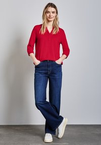 Rode blouse met lange mouwen, V-hals en knoopdetails, gecombineerd met donkerblauwe flared jeans en witte sneakers op een grijze achtergrond.
