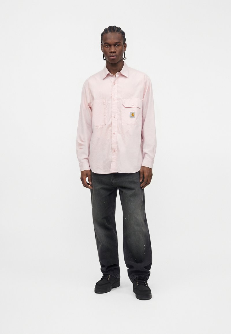 Man standing wearing a light pink long-sleeve button-up shirt with pockets and dark gray loose jeans against a plain white background.