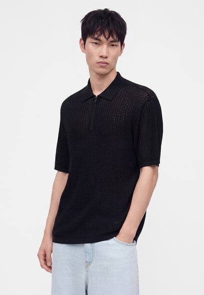 Jeune homme aux cheveux courts et noirs portant un polo noir texturé à manches courtes et un jean bleu clair sur un fond uni.