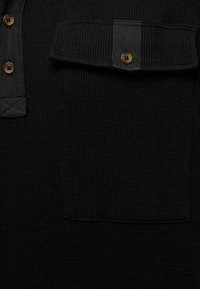Chemise noire texturée avec un design à col, comportant une poche et des boutons marron. Le tissu présente un motif en relief, semblable à un gaufret.