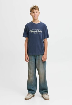 Camiseta azul marino con texto blanco, jeans descoloridos de corte relajado y zapatos beige con cordones y suelas de goma, de pie contra un fondo liso.