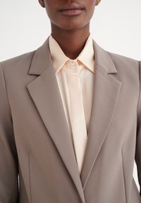 Blazer beige con rever affilati e tessuto testurizzato, indossato sopra una camicia a bottoni pesca chiaro con finitura liscia e colletto a punta.
