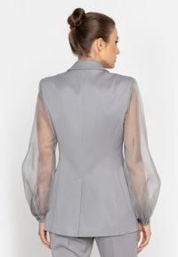 Szary żakiet z fasonem dopasowanym, z przezroczystymi bufkami, usztywnionymi ramionami oraz wcięciem w talii. Gładki materiał z delikatnym połyskiem, z mankietami zapinanymi na guziki.