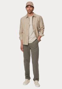 Beige leichte Jacke mit Taschen, kombiniert mit einem weißen T-Shirt, olivgrünen Chinos und beigen Sneakers. Das Modell trägt eine passende Mütze.