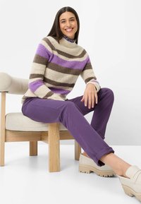 Gestreepte trui in crème, bruin en paarse fleece, gecombineerd met paarse broek. Zittend op een houten stoel, met beige plateau schoenen.