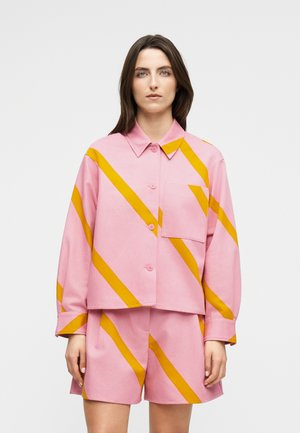 Femme portant une chemise rose à boutons et un short assorti avec des rayures diagonales orange vives sur un fond uni.