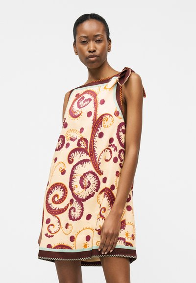Γυναίκα που φοράει αμάνικο φόρεμα μέχρι το γόνατο με σχέδια παϊλέι σε μπορντό και πορτοκαλί σε εκρού φόντο και λεπτομέρεια δέσιμου στον ώμο.