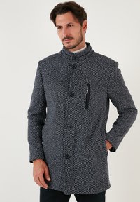 Cappotto da uomo in navy con un motivo tessuto testurizzato. Presenta un colletto alto, bottoni frontali e una tasca con zip sul lato sinistro del petto.