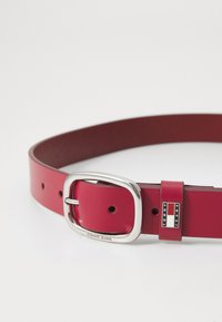 Red synthetic belt with a shiny silver rectangular buckle, featuring a logo accent and multiple adjustment holes along the strap.