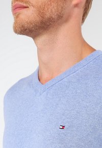 Pull léger à col en V bleu clair en tissu tricoté doux, avec une petite étiquette logo sur le côté gauche de la poitrine.