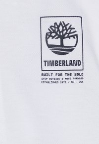 Tessuto bianco con un logo Timberland nero che presenta un design ad albero, circondato da un bordo rettangolare e testo sottostante che indica i dettagli del marchio.