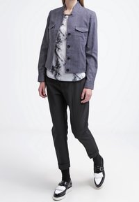Giacca grigia aderente con colletto rialzato, due tasche frontali, abbinata a una camicetta bianca con motivo floreale nero e pantaloni neri a sigaretta.