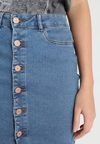 Džínová sukně ve středně modré barvě, s předním zapínáním na knoflíky měděné barvy a úzkým střihem s kapsami.
