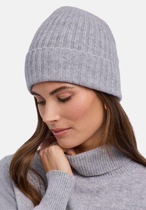 Kobieta w jasnoszarej prążkowanej dzianinowej czapce i pasującym golfie, spoglądająca w dół z ręką przy brodzie.