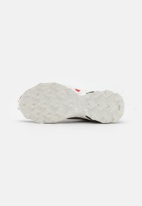 Suela de zapatilla de running blanca con patrones de agarre texturizados, material de goma y una ligera curvatura. Incluye un acento rojo cerca del talón.