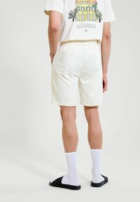 Persona vestida con pantalones cortos blancos, camiseta gráfica blanca, calcetines blancos y sandalias negras de tipo slide, de pie con las manos en los bolsillos, de espaldas a la cámara.