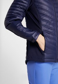 Marinblå jacka med vadderade, quiltade ärmar och en slät kropp. Dragkedjeficka på sidan. Materialet ser lätt och flexibelt ut.