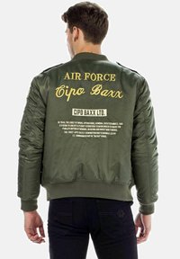 Olijfgroene bomberjack met gouden tekst op de achterkant; voorzien van ribgebreide manchetten en zoom; gepresteerde branding en historische tekstdetails.