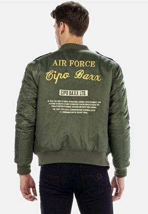 Olivgrünes Bomberjackett mit goldener Schrift auf dem Rücken; ausgestattet mit gerippten Bündchen und Saum; geprägt mit Branding und historischen Textelementen.